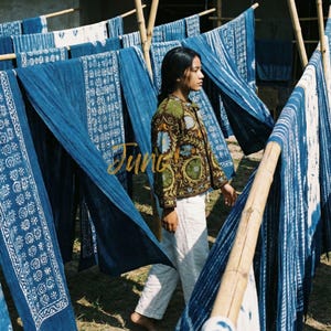May include: A woman in a brown jacket with floral embroidery and white pants walks past indigo-dyed textiles hanging on bamboo poles. The textiles have white patterns. The word "Jung" is written in gold script.