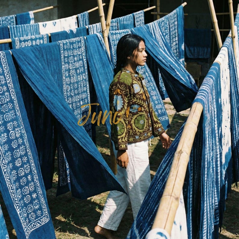 May include: A woman in a brown jacket with floral embroidery and white pants walks past indigo-dyed textiles hanging on bamboo poles. The textiles have white patterns. The word "Jung" is written in gold script.