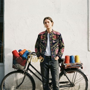 Könnte beinhalten: Eine Person trägt eine marineblaue Jacke mit farbenfrohen Blumenstickereien und steht neben einem schwarzen Fahrrad mit einem Korb voller Garn. Die Jacke hat rosa, grüne und goldene Akzente. Das Fahrrad hat Garnspulen im Korb.