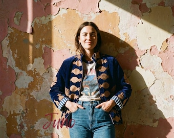 May include: A dark blue velvet jacket featuring a repeating gold pomegranate pattern, worn over a graphic t-shirt and blue denim jeans. The jacket has long sleeves with striped cuffs and a front button closure, paired with classic blue denim pants.