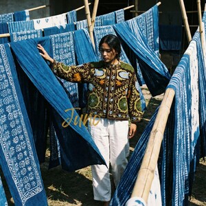 May include: A woman stands among indigo-dyed textiles hanging on bamboo racks. She wears a brown jacket with floral embroidery and white pants. The textiles feature various white patterns on a deep blue background. The word "Juno" is written in gold script.