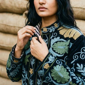 Op de afbeelding: Zwarte geborduurde jas met bloemen- en bladerpatronen in groen, goud en blauw. De jas heeft een hoge kraag en wordt dichtgeknoopt. De achtergrond is een neutrale bruine kleur.