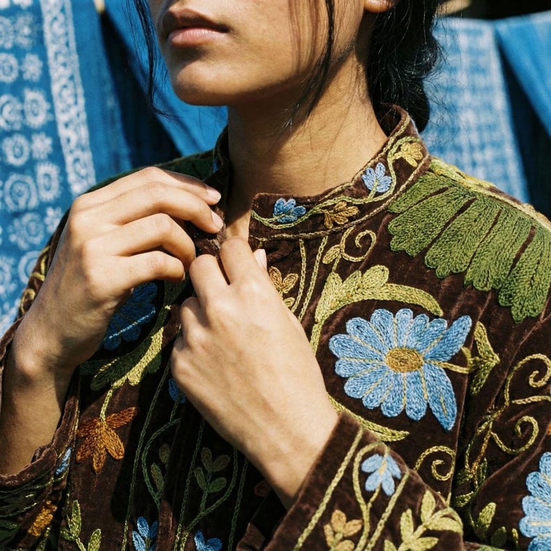 May include: A brown velvet jacket with intricate floral embroidery in shades of blue, green, and gold. The jacket features a mandarin collar and detailed stitching, showcasing a bohemian style.