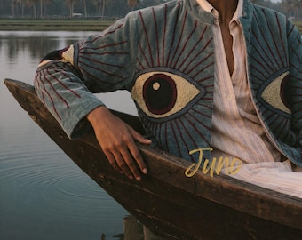 May include: A blue-grey quilted jacket featuring large embroidered eye motifs on the chest and sleeves, with radiating red and gold stitching. The jacket is worn over a light-colored, vertically striped button-down shirt with the word Juno in gold.