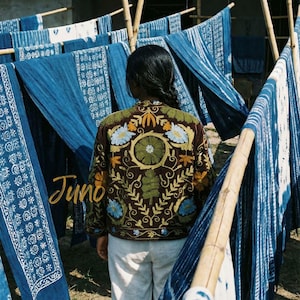 May include: A brown jacket with colorful floral embroidery, the word "Juno" in gold script, and white pants. Indigo-dyed textiles hang on bamboo poles in the background. The jacket features blue, green, and yellow floral designs.