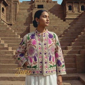 May include: Cream-coloured jacket with detailed floral embroidery in purple, pink, and green. The jacket features a mandarin collar and long sleeves. The word "Juno" is written in gold script at the bottom. The jacket is worn over a white dress.