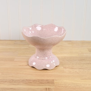 May include: A pink ceramic pedestal bowl with a speckled finish. The bowl has a scalloped rim and base, and is elevated on a short, rounded pedestal. The bowl is likely used for serving desserts or displaying decorative items.