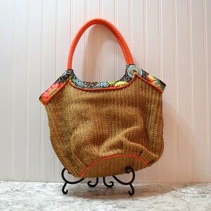 May include: A brown woven tote bag with a bright orange trim and a colorful floral fabric lining. The bag is on a black metal stand.