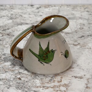 May include: A small, white ceramic pitcher with a brown handle and green and brown bird and butterfly designs.
