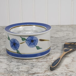 May include: A white ceramic butter dish with a blue and gold trim and a floral design. The dish has a lid and is sitting on a light gray countertop next to a wooden spatula.