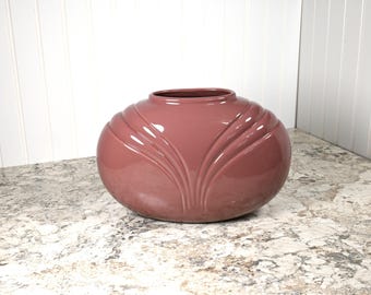 May include: A round, dusty rose-colored ceramic vase with a wide opening. The vase features a series of vertical, curved ridges that create a petal-like design. The vase is sitting on a speckled countertop.
