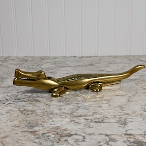 May include: A gold-colored alligator nutcracker. The alligator's mouth is open and its teeth are visible. The nutcracker is made of metal and has a detailed design.