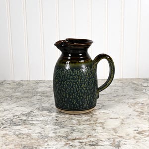 May include: A green and brown ceramic pitcher with a speckled glaze. The pitcher has a rounded body and a handle.