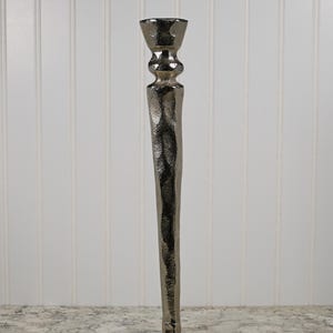 May include: A silver metal candle holder with a textured, hammered finish. The holder has a tall, slender design with a wide, open top.