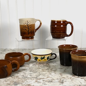 May include: Six ceramic mugs in various styles and colors. Two mugs have a brown and white tree design, one has a brown and white floral design, one has a brown and white striped design, and two have a brown and black ombre design.