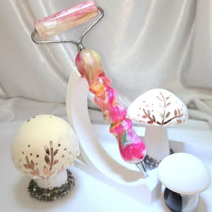 May include: A facial roller with a swirled pink, yellow, and white resin handle and a matching roller head, supported by a white curved stand. Two decorative mushroom figures with gold leaf accents and a small black and white mushroom are present.