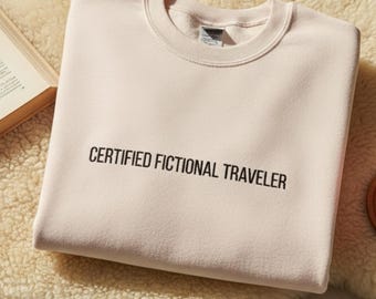 May include: A light pink sweatshirt with the text "CERTIFIED FICTIONAL TRAVELER" in black. The sweatshirt is folded on a fluffy, cream-colored rug. A book, glasses, and a cup of coffee are also visible.