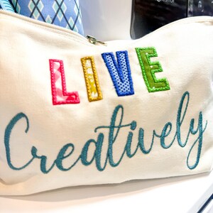 May include: Cream-colored canvas zippered pouch with the embroidered words "LIVE Creatively" in colorful, stitched letters. The pouch is partially open, revealing a glimpse of fabric and a sewing machine in the background.