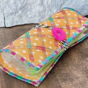 May include: A yellow fabric roll-up organizer featuring a white polka dot pattern, secured with a bright pink button and a black and white striped cord. The edges are finished with decorative multi-colored zigzag stitching throughout.