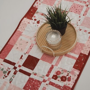 May include: A patchwork table runner in red, pink, and white. The design includes hearts, roses, and gingham patterns. A round bamboo tray holds a small potted plant and a glass candle holder. The runner is suitable for a table.