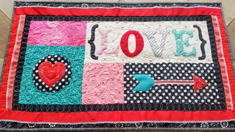 May include: A quilted wall hanging with the word "LOVE" in bold letters. The design incorporates pink, teal, and black polka dot patterns, with a red heart and arrow accents. The border is red with a black and white trim.