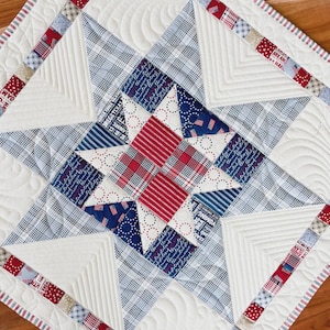 May include: A square quilt featuring a central star pattern composed of red, white, and blue fabrics. The design includes plaid, striped, and text-printed textiles with intricate white machine-quilted feather and geometric line patterns.