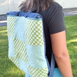 Op de afbeelding: Een gewatteerde rugzak met een patchwork-ontwerp. De tas heeft een combinatie van lichtblauwe en groene ruitjesstof en effen lichtblauwe stof. De bandjes en de afwerking zijn van effen blauwe denim. De tas wordt over de schouder gedragen.