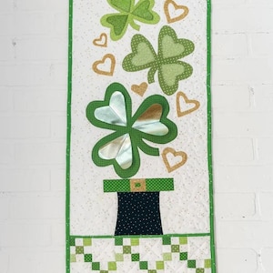 May include: A green and gold quilted wall hanging with a leprechaun's hat, shamrocks, and hearts. The background is white with green polka dots.