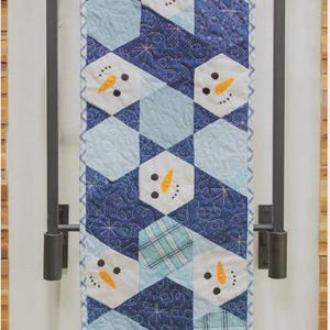 May include: A quilted table runner with a repeating pattern of snowman faces on a blue and white geometric design. The snowmen have orange carrot noses and black dot eyes. The runner is displayed on a dark metal rack.