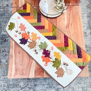 May include: A white table runner with a brown border features a design of autumn leaves and acorns. The runner is decorated with a variety of autumn colours, including orange, yellow, red, and green. The runner is perfect for adding a touch of autumn charm to your home decor.