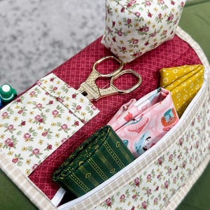May include: A floral print sewing organizer with a red quilted base. It features a pin cushion, gold scissors, and pockets holding folded fabric in various patterns and colors. The organizer is designed for sewing and crafting.