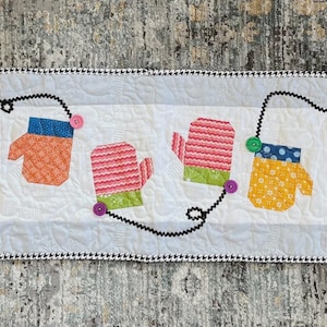 May include: A quilted table runner featuring colorful mittens in various patterns and colors, including green polka dots, orange, pink stripes, and yellow with blue polka dots. The mittens are connected by a black dotted line, with a black and white checkered border.