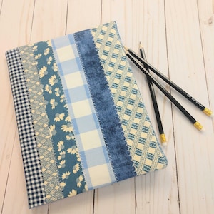 Pode incluir: Um caderno coberto de tecido com um design de patchwork em tons de azul e branco, com padrões xadrez, florais e geométricos. Três lápis pretos com pontas douradas estão sobre o caderno.