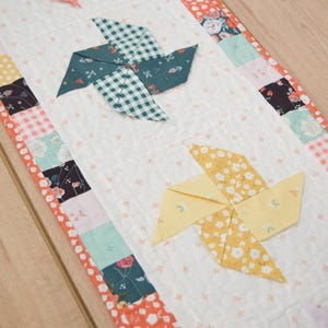 May include: A quilted table runner with a white background and colorful patchwork borders. The runner features three origami-style pinwheel designs in shades of pink, green, and yellow, adding a whimsical touch to the home decor.