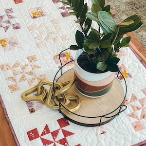 Puede incluir: Una decoración de mesa con un edredón blanco con patrones florales y geométricos. Una bandeja redonda de madera con borde de metal negro sostiene una planta en maceta. Un nudo decorativo dorado se encuentra sobre el edredón.