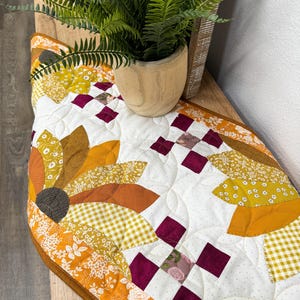 Könnte beinhalten: Ein gesteppter Tischläufer mit einem Sonnenblumenmuster in Orange-, Gelb- und Brauntönen. Der Läufer hat einen weißen Hintergrund mit einem burgunderfarbenen Rand.