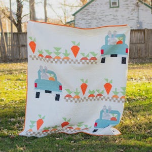 Puede incluir: Un edredón blanco con un borde naranja, que presenta un patrón repetido de camiones azul claro que transportan zanahorias y pasajeros conejos. El diseño incluye zanahorias naranjas y copas verdes, con un patrón de cuadros.