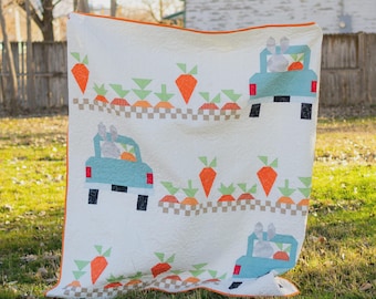 Könnte beinhalten: Eine weiße Steppdecke mit orangefarbenem Rand, die ein sich wiederholendes Muster aus hellblauen Lastwagen mit Karotten und Hasenpassagieren zeigt. Das Design umfasst orangefarbene Karotten und grüne Spitzen mit einem Schachbrettmuster.