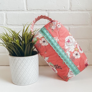 May include: A quilted, rectangular bag with a floral pattern in shades of pink, white, and green. The bag features a mint green zipper and lace trim. A braided handle in pink and white completes the design. A potted plant is next to the bag.