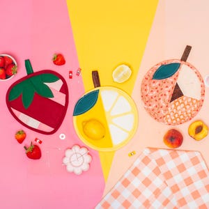 Può includere: Tre presine in tessuto a forma di fragola, fetta di limone e pesca, disposte su uno sfondo multicolore. La fragola è rossa e verde, il limone è giallo e bianco, e la pesca presenta tonalità arancioni e color crema.