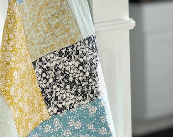 Op de afbeelding: Een doorgestikte deken met een patchworkontwerp van diverse bloemenpatronen in tinten mosterdgeel, teal, zwart en wit. De stoffen vierkanten zijn aan elkaar genaaid met zichtbare stiklijnen, wat zorgt voor een gestructureerd oppervlak.