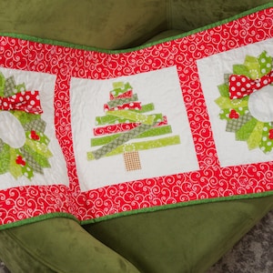 May include: A festive Christmas table runner featuring three white squares with holiday designs. The squares include two wreaths with red bows and a Christmas tree. The runner has a red and white swirl border and green trim.