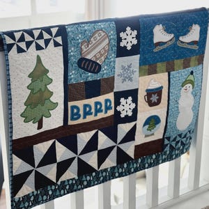 May include: A winter-themed quilt with various appliqued designs, including a Christmas tree, snowflakes, a mitten, ice skates, a snowman, and a hot beverage. The word "BRRR" is stitched on the quilt. The quilt is displayed on a wooden railing.