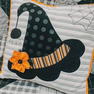May include: A square throw pillow featuring a grey and white striped background with a black polka-dot witch hat appliqué. The hat includes an orange and black striped band, a fabric orange flower, and a dangling black spider with stitched legs.
