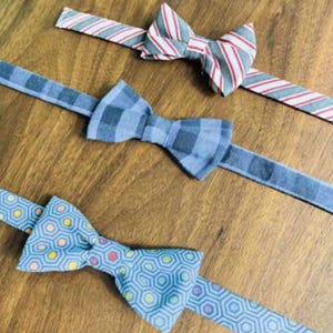 Könnte beinhalten: Drei Fliegen mit verschiedenen Mustern werden auf einer Holzoberfläche gezeigt. Eine Fliege hat ein blau-weißes geometrisches Muster, eine andere ein blaues Karomuster und die dritte rot-weiße Streifen.