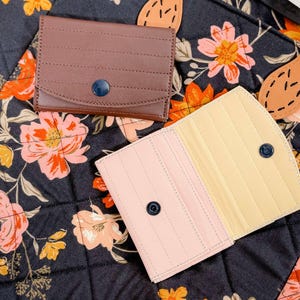 May include: A brown leather wallet with a blue button closure sits on a floral patterned fabric. A pink and yellow wallet is open, revealing its interior. The background features a dark floral pattern with orange and yellow flowers.