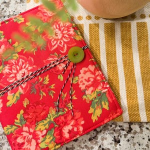 May include: A red floral patterned journal cover with a green button closure and black and white twine. The journal is next to a yellow and white striped fabric.