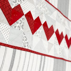 May include: A close-up of a quilt with a repeating heart pattern in red and white. The quilt features various geometric shapes and patterns, including stripes and floral designs. The border is a solid red.