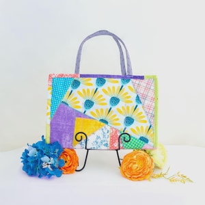 May include: A colorful patchwork tote bag with a floral design, featuring yellow daisies on a white background. The bag has a purple handle and is displayed on a black metal stand, with blue and orange flowers in front.