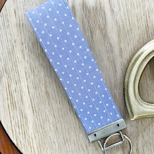 Puede incluir: Una pulsera de tela azul claro para llaves con un patrón de cruces blancas. La pulsera tiene un cierre de metal plateado y un llavero plateado. La pulsera está sobre una superficie de madera.